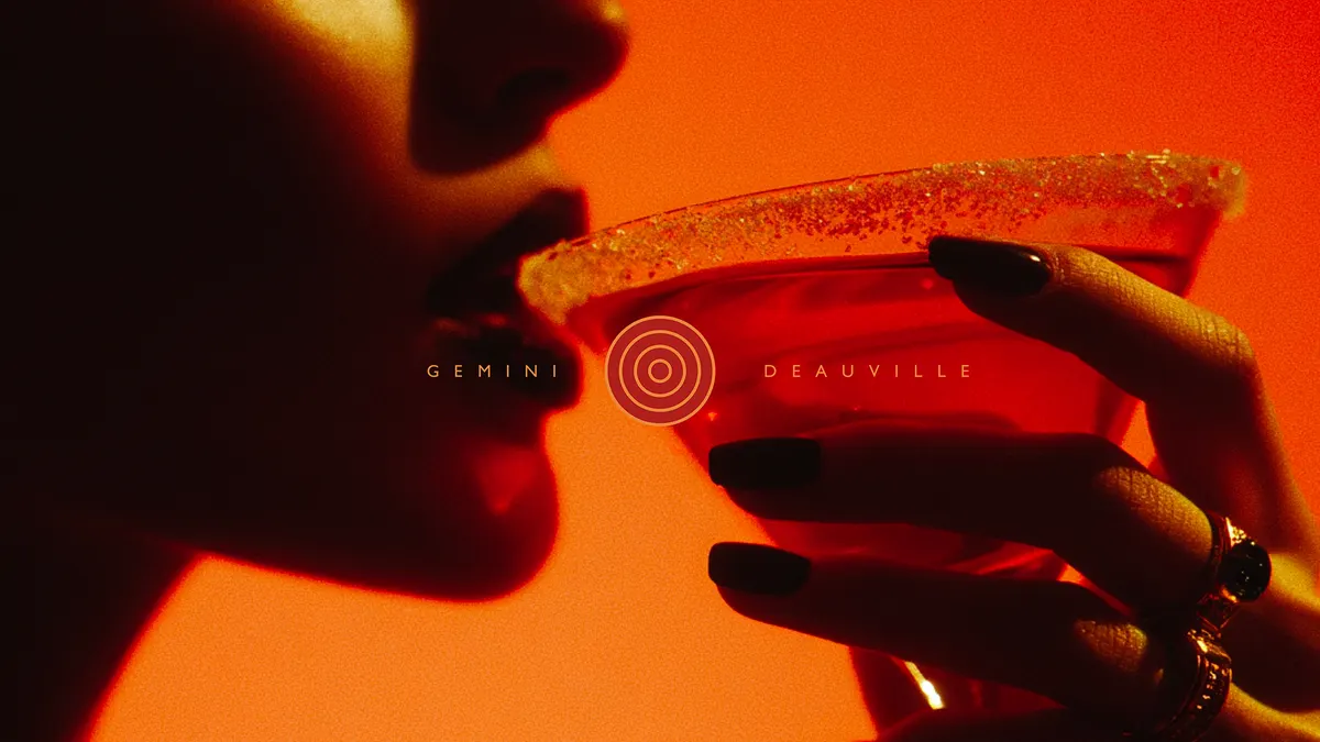 Femme buvant un cocktail rouge marqué Gemini Deauville, lumière orangée