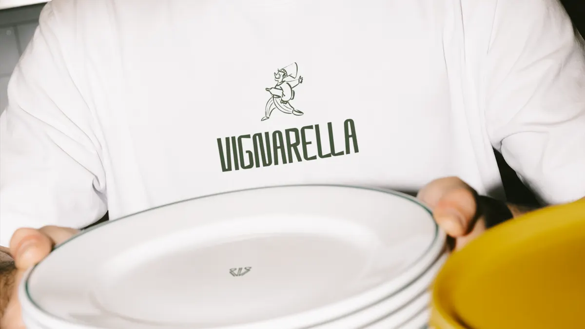 Serveur en veste blanche brodée du logotype Vignarella et d'un personnage illustré, tenant une pile d'assiettes